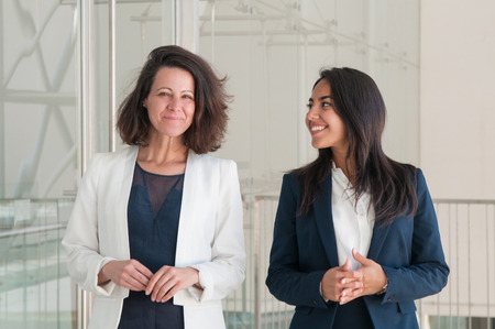 Two joyful beautiful business women in office hall. Colleagues looking at camera and standing in office hall. Business women portrait concept. Front view.の写真素材