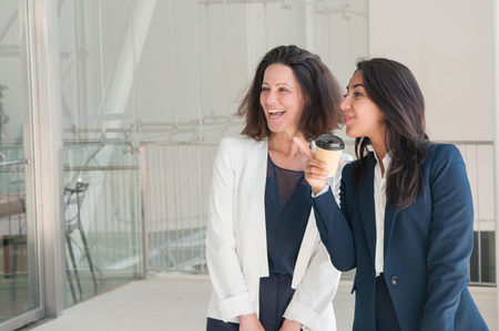 Two positive colleagues relaxing in office hall. Attractive women standing, pointing away and drinking coffee. Break and relaxation concept.の写真素材
