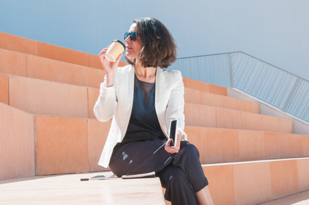 Successful businesswoman enjoying tasty coffee during break outside. Woman in sunglasses and white jacket holding tablet and drinking coffee from paper cup. Coffee break conceptの写真素材