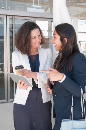 Two excited business women pointing at tablet screen in hall. Colleagues or partners standing with tablet computer. Technology and news concept.の写真素材