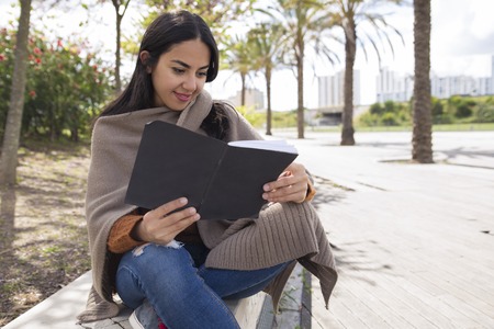 Smiling pretty woman reading copybook and studying outdoors. Young woman sitting on parapet with blurred trees and walkway in background. Education concept.の写真素材