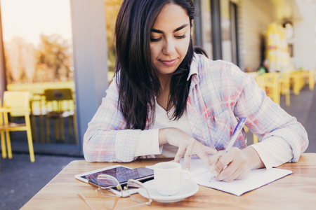 Serious woman making notes in outdoor cafe. Pretty young lady wearing casual clothes and sitting at table with chairs and building in background. Education concept. Front view.の写真素材