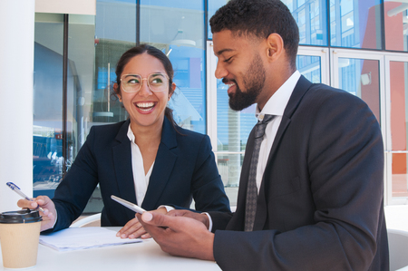 Happy colleagues working and using tablet in outdoor cafe. Business man and woman wearing formal clothes and sitting with building glass wall in background. Business work concept.の写真素材