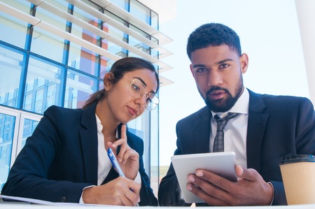 Serious business people working and using tablet outdoors. Business man and woman wearing formal clothes and sitting with building glass wall in background. Business and technology concept. Front viewの写真素材