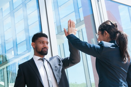 Positive business people high fiving outdoors. Business man and woman wearing formal clothes and standing with building glass wall in background. Unity concept.の写真素材