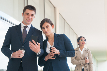 Positive and sly coworkers enjoying short work break. Business man and woman holding cells, coffee cup and looking at camera, their colleague walking to office behind them. Communication conceptの写真素材