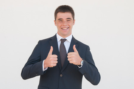 Cheerful recent graduate or intern happy to start his career. Young man in office jacket and tie showing thumb up with both hands and smiling at camera. Approval conceptの写真素材