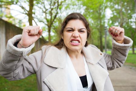 Angry frustrated girl learning bad news. Young woman in casual jacket walking outdoors and raising arms with clenched fists in furious gesture. Rage or negative emotions conceptの写真素材