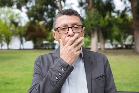 Shocked middle-aged man covering mouth with hand in park. Guy wearing casual clothes and standing with green lawn and trees in background. Shock and nature concept. Front view.の写真素材