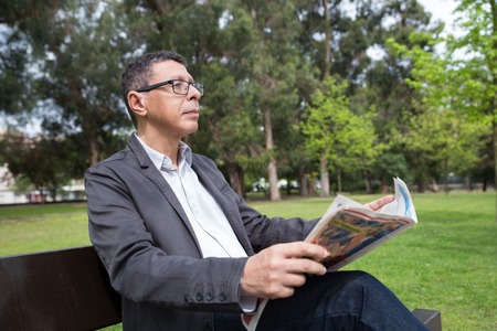 Relaxed man reading newspaper and sitting on bench in park. Guy wearing casual clothes and sitting with green lawn and trees in background. News and nature concept.の写真素材