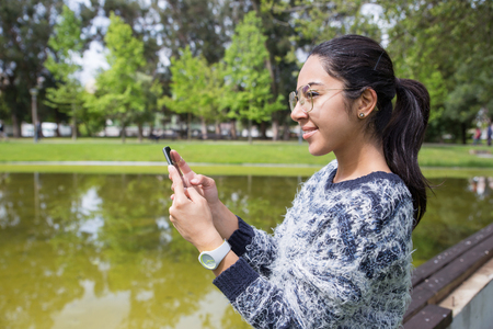 Smiling young woman using smartphone in park. Pretty lady wearing sweater and standing with pond and green trees in background. Communication and nature concept. Side view.の写真素材