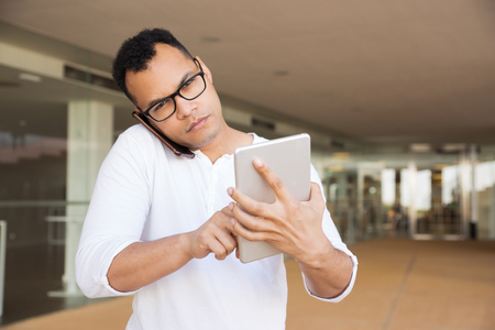 Medium shot of serious young mixed-race man in spectacles and white T-shirt standing at office building, working on tablet, talking on phone, looking at camera. Front view. Lifestyle conceptの写真素材