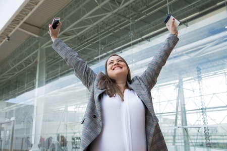 Cheerful business woman raising arms outdoors. Lady holding plastic cup, smartphone and standing with building glass wall in background. Break and success concept. Front view.の写真素材