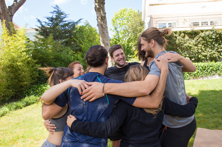 Cheerful men and women hugging each other after workout in park. United team people in fitness apparels forming circle and hugging outdoors. Unity and collaboration conceptの写真素材