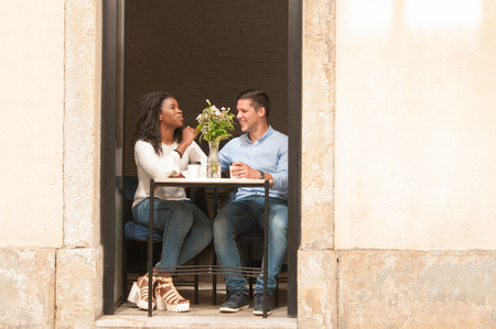 Positive handsome Caucasian guy dating beautiful Afro American girl. Happy interracial couple sitting at table in doorway and drinking coffee. Romantic date conceptの写真素材