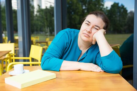 Bored woman spending time at outdoor cafe. Pensive woman leaning on her arms and looking ahead after...