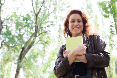 Portrait of happy woman with notepad. Cheerful female university student standing in park with small...