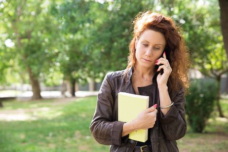 Serious young woman talking on phone. Pensive pretty brunette walking on campus with book and making phone call. Communication conceptの写真素材