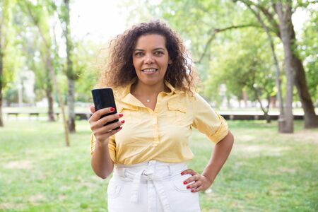 Smiling woman reading news on smartphone in summer park. Young lady wearing casual shirt and skirt...