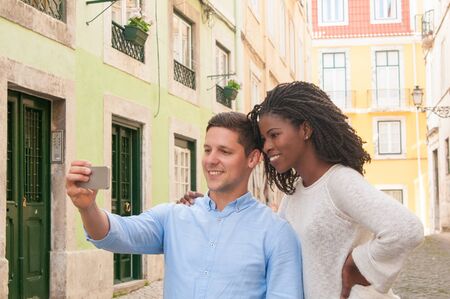 Cheerful multi-ethnic couple posing for self-portrait on smartphone. Smiling attractive black woman embracing boyfriend while they taking selfie. Tourists conceptの写真素材