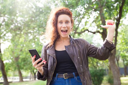 Cheerful lady showing thumb up and holding smartphone in park. Young woman holding gadget, screaming and standing with blurred green trees in background. Success or good news concept. Front view.の写真素材
