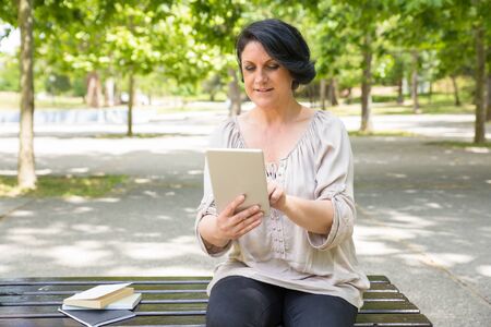 Peaceful middle-aged woman reading on tablet screen. Focused Caucasian lady sitting near books and notes on park bench and using digital device. Online learning conceptの写真素材