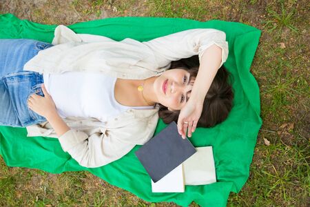 Peaceful carefree college girl happy to finish study book. Top view of young woman lying down on grass near textbooks and notebooks and smiling at camera. Relaxing conceptの写真素材