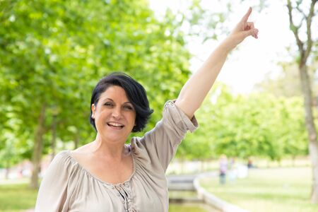 Joyful excited woman standing outdoors against green trees and pointing finger up. Middle aged black haired lady raising hand and smiling at camera. Pointing up conceptの写真素材