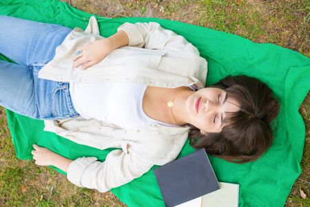 Peaceful tranquil girl relaxing in park. Top view of young woman lying on grass with closed eyes....