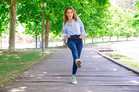 Carefree young woman in casual outfit walking straight leg raise. Positive girl with wavy hair strolling in park. Playful girl conceptの写真素材