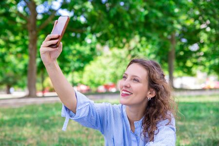 Smiling young woman choosing angle for self-portrait. Happy curly-haired girl photographing herself...