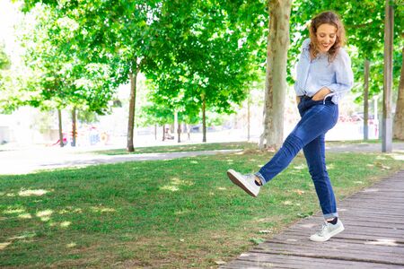 Positive woman ambling in park. Cheerful curly-haired girl in jeans walking straight leg raise...