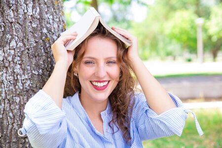Jolly girl with book above head preparing for exam in park. Portrait of happy young woman in casual shirt resting in park. Education conceptの写真素材