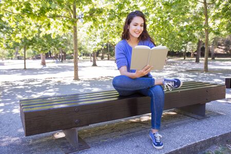 Positive curious woman in casual clothing reading book in park. Cheerful excited girl enjoying literature. Leisure conceptの写真素材
