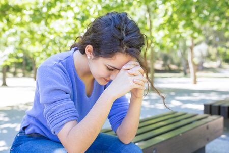 Upset young woman sitting on bench in city park. Worried girl leaning on knees while focusing on thoughts. Problems conceptの写真素材