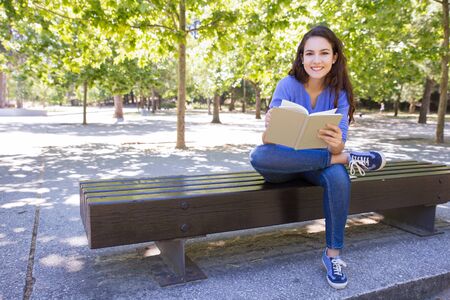 Happy student girl enjoying reading in summer park. Cheerful woman sitting on bench in park. Hobby conceptの写真素材