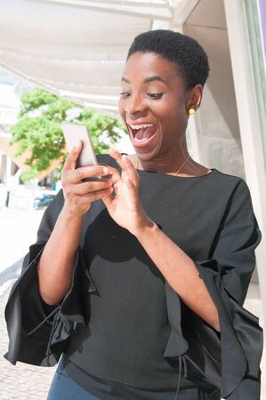 Amazed joyful African American woman getting good news from phone message. Happy excited black lady using smartphone, looking at screen and shouting with joy. Phone using or amazement conceptの写真素材