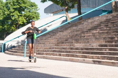 Cheerful excited girl riding scooter near building and stairs. Short haired Afro American rider...