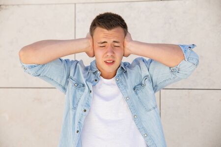 Stressed young man covering ears with hands at wall outdoors. Handsome white guy wearing jeans...