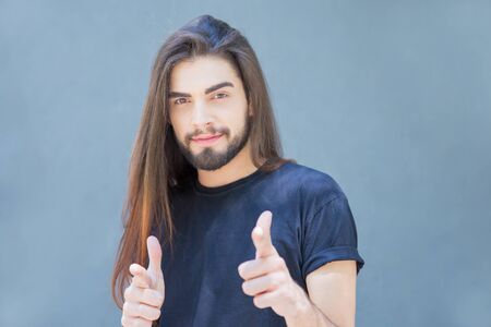 Confident smiling Latin young man pointing with finger guns. Positive attractive guy with long...