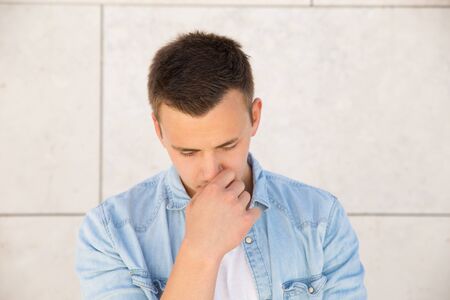Pensive young man touching face at wall outdoors. Handsome white guy wearing jeans shirt and...