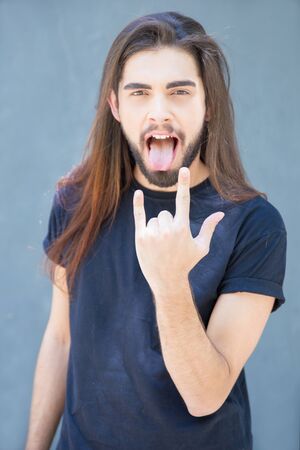 Handsome cool band player showing rock gesture. Latin young man sticking out tongue and making devil...