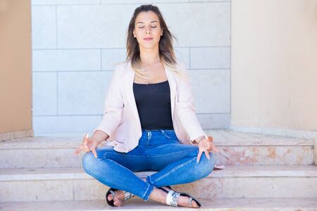Peaceful tranquil young woman meditating near city building. Serene beautiful woman sitting on stair in lotus position, making zen gesture with fingers. Yoga or stress relief conceptの写真素材