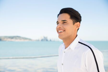 Happy young Latin man at seaside. Handsome man wearing shirt and standing with sea and blue sky in background. Tourism concept.の写真素材