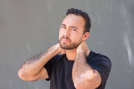 Young man touching neck and looking at camera. Unhappy bearded man holding hands at neck on grey...