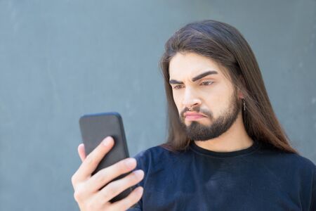 Upset bearded brunet with piercing in nose looking at smartphone. Handsome young man holding modern phone. Technology conceptの写真素材
