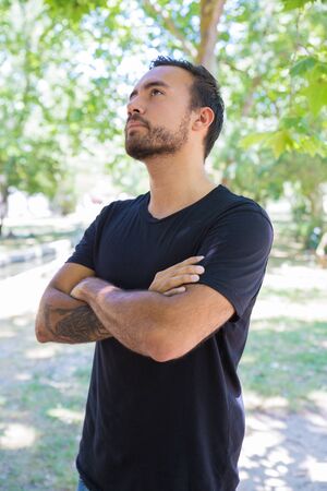Pensive man with crossed arms looking up. Handsome bearded young man in black t-shirt standing outdoor. Gesture conceptの写真素材