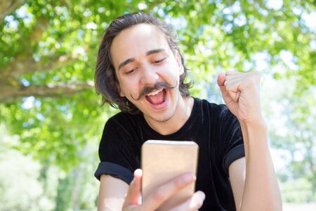 Happy young man looking at smartphone in park. Closeup shot of smiling stylish guy with raised fist. Technology conceptの写真素材