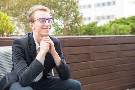 Cheerful manager enjoying work break outside. Happy young business man in eyeglasses and jacket sitting on bench, leaning chin on hands, looking away and smiling. Business portrait conceptの写真素材