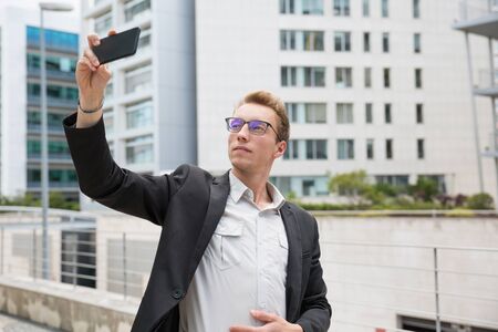 Pensive calm businessman taking selfie. Young man in office suit and eyeglasses using phone camera...